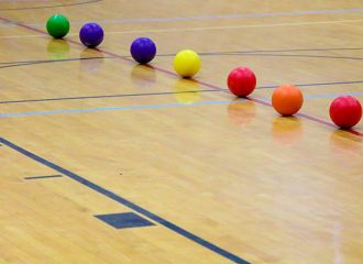 Indoor Games (Dodgeball, Rounders) Dodgeballs lined up in the center of a gymnasium waiting for the start of the game. The seven balls are red, orange, yellow, blue and green. Dodgeball is an indoor team sport.